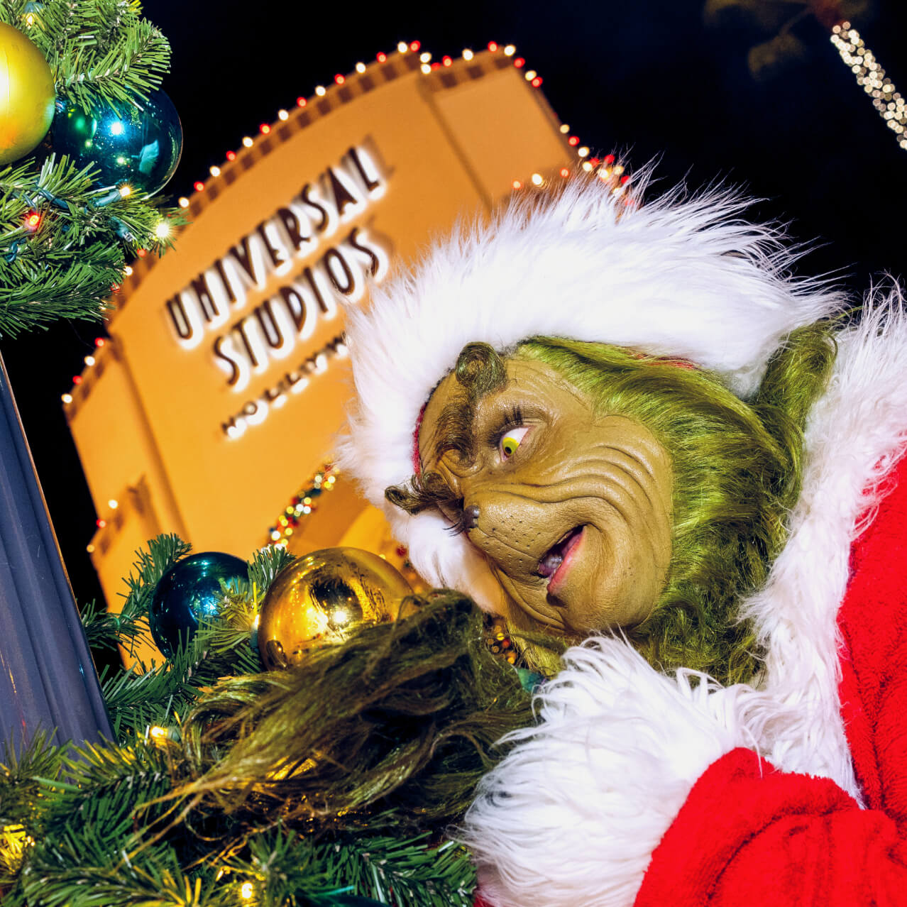 The Grinch in Santa gear closely inspecting a gold ornament on garland wrapped around a pole at Universal Studios Hollywood.
