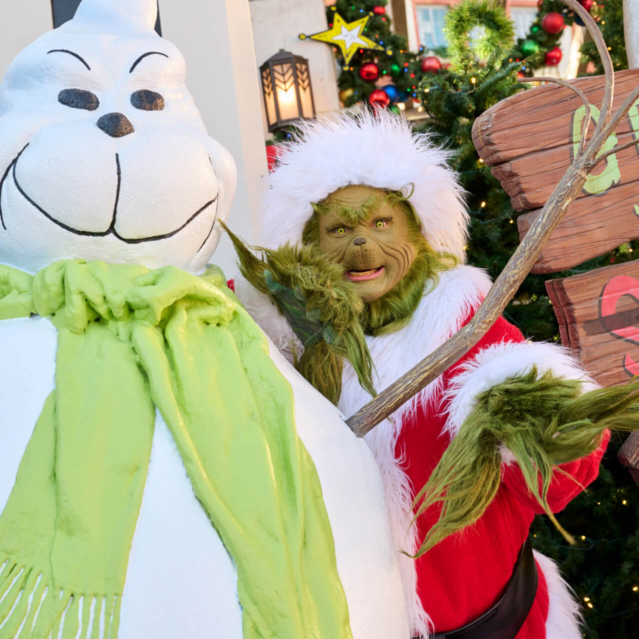The Grinch, in his Santa hat and coat, posing mischievously with a green-scarved snowman that looks a lot like him.