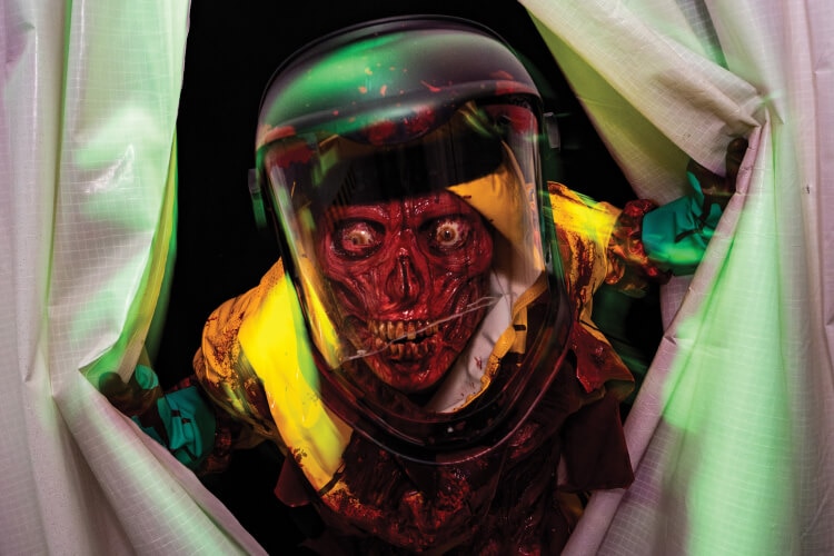 A zombie scareactor with a deteriorated face in bloody hazmat suit lunging through plastic curtains.
