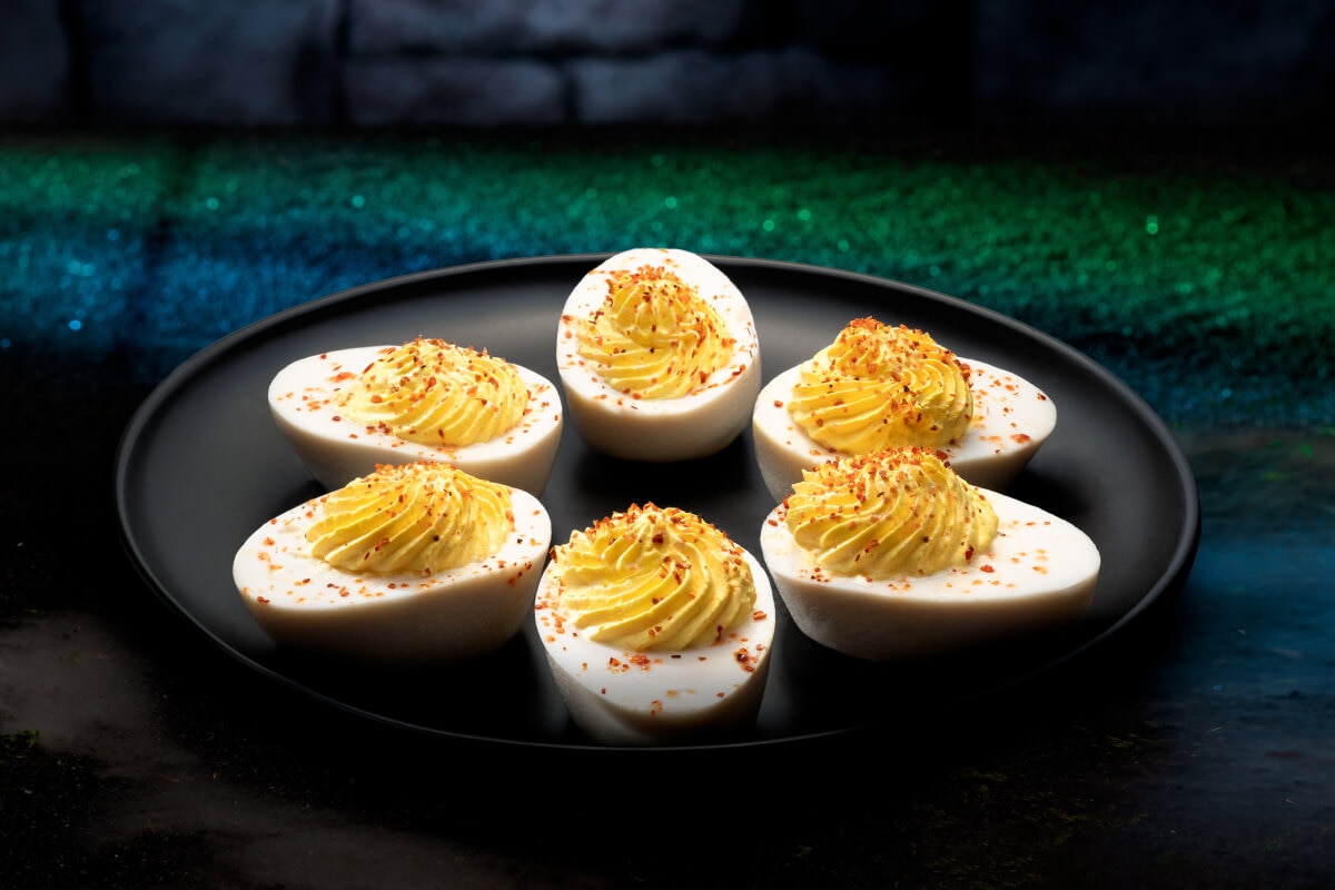 A black plate of five deviled eggs with red flakes of seasoning against a green carpet and dark stone wall background.