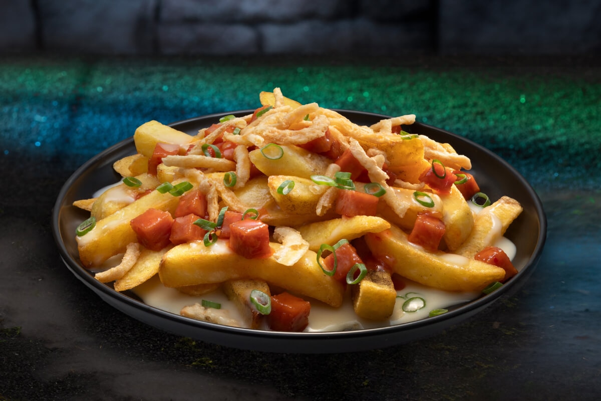 A black plate of fries topped with vegetables, cheese and other items against a green-lit floor and dark stone wall background.