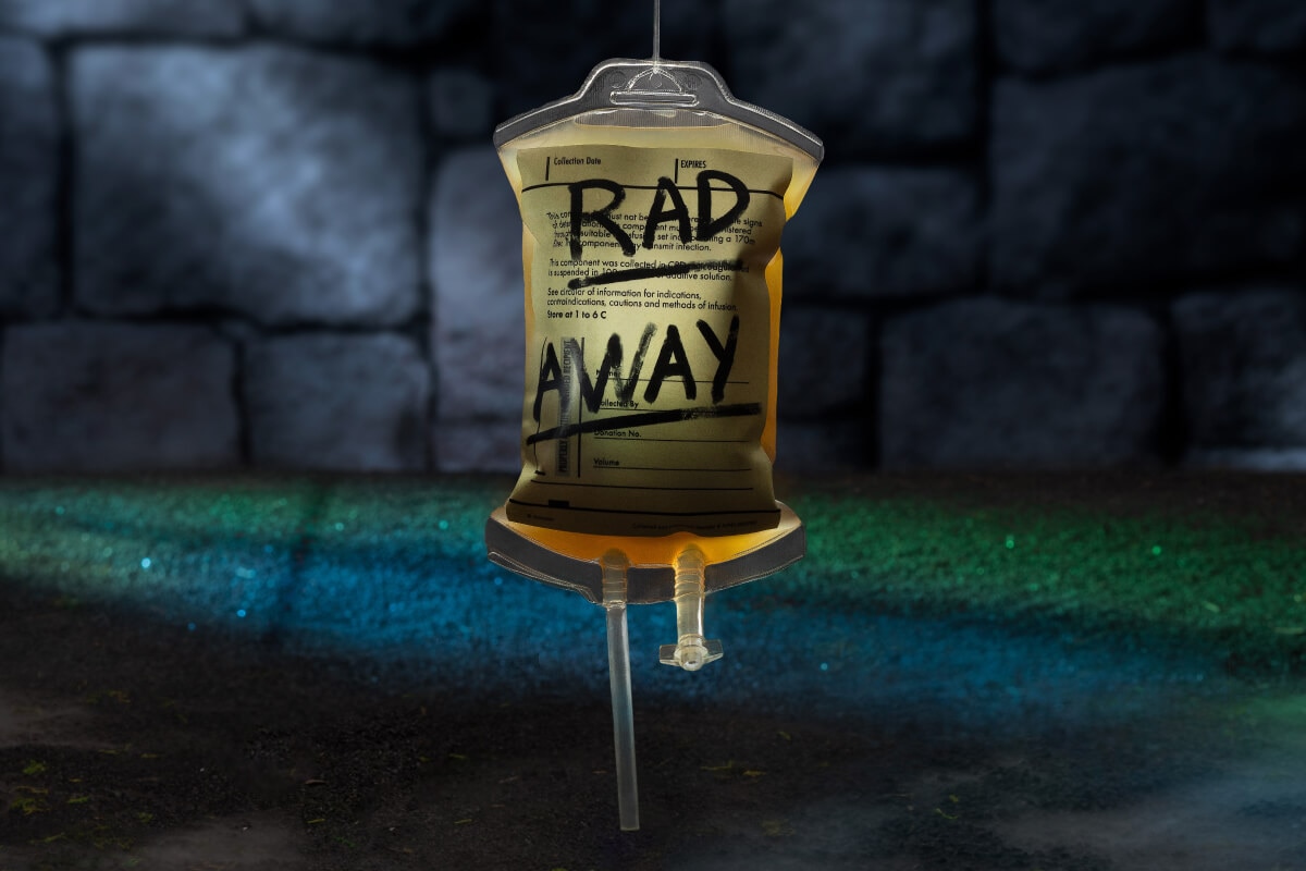 An IV bag of amber-colored liquid with the words “RAD AWAY” in marker against a green carpet and dark stone wall background.