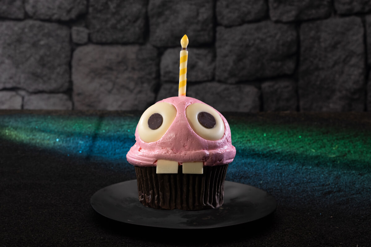 Cupcake of Five Nights at Freddy’s treat with eyes, teeth, red frosting and candle against a green carpet and stone wall.