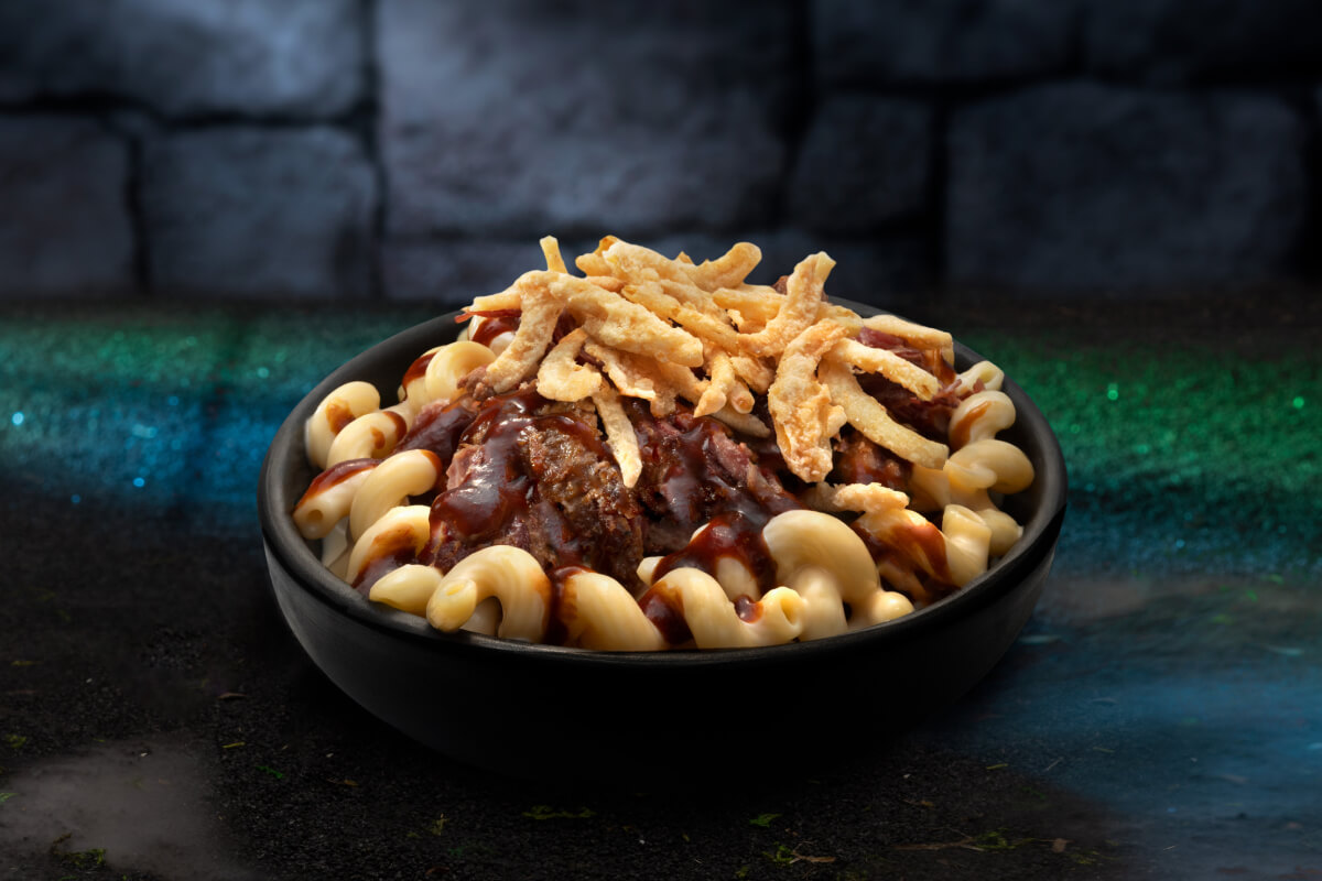 A black bowl of mac and cheese topped with brisket, sauce and fries against a green carpet and dark stone wall background.