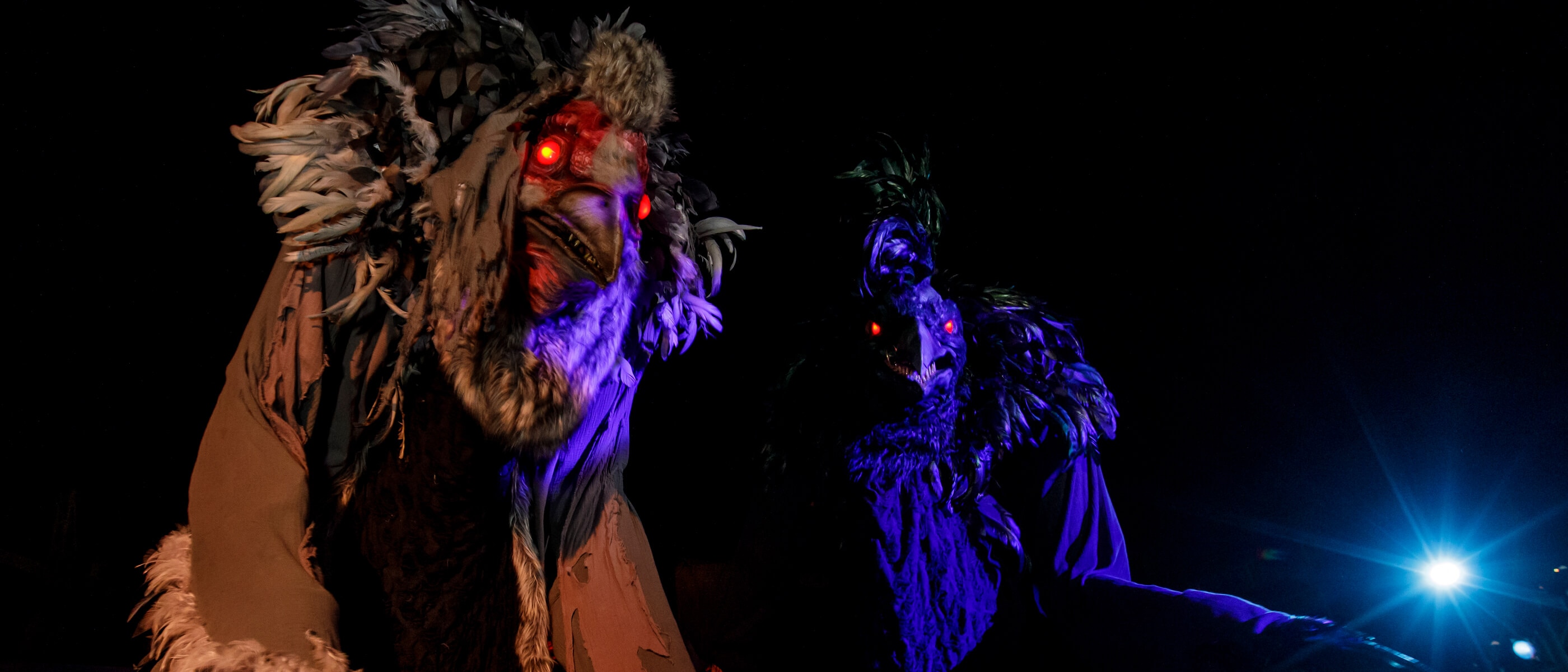 A pair of towering scareactors with crow feathers, beaks, and glowing red eyes walking the night streets.