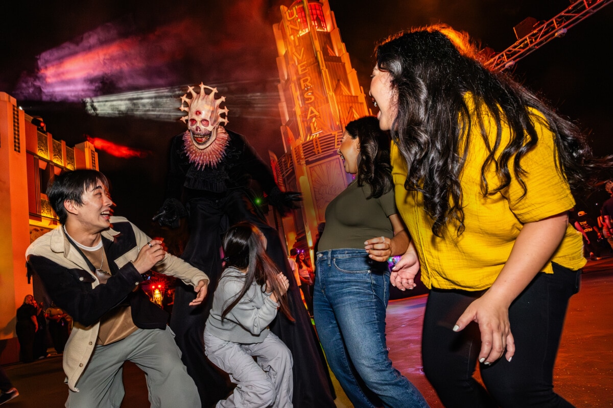A tall scareactor with a skeletal face, three red eyes, and a halo of horns terrifying a group of four friends in the night streets.