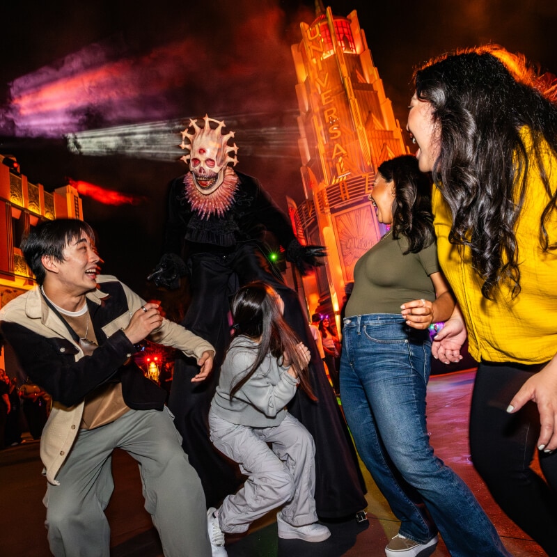 A tall scareactor with a skeletal face, three red eyes, and a halo of horns terrifying a group of four friends in the night streets.