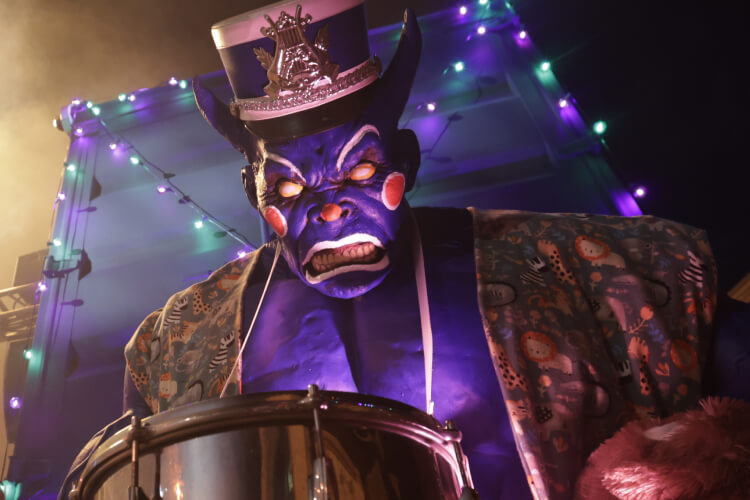A giant purple figure with long ears, blank white eyes, and a band uniform hat snarling menacingly behind a large silver drum.