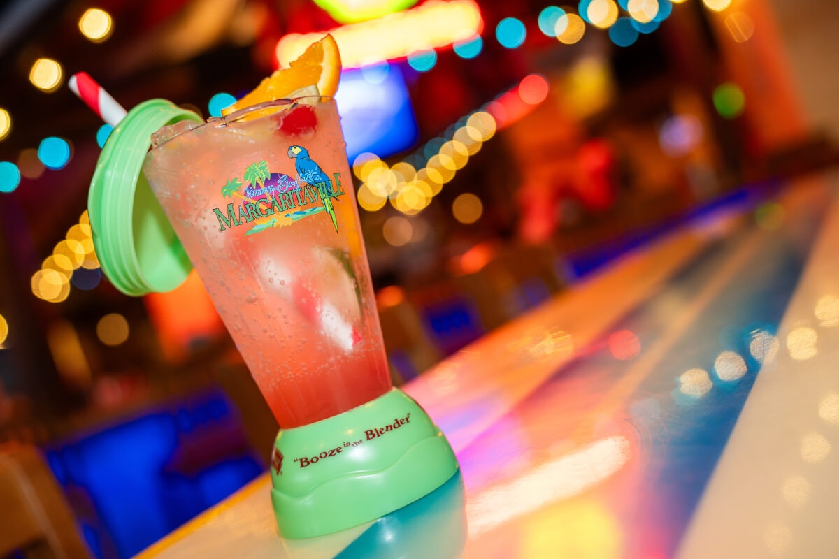 An orange-red drink with fruit in a blender-shaped glass with “MARGARITAVILLE” and “Booze in the Blender” on a wooden table.