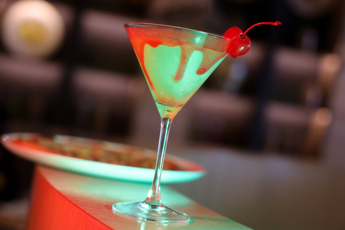 A tall martini glass containing a green drink with red drippings and a cherry in front of a food plate on a curved counter.