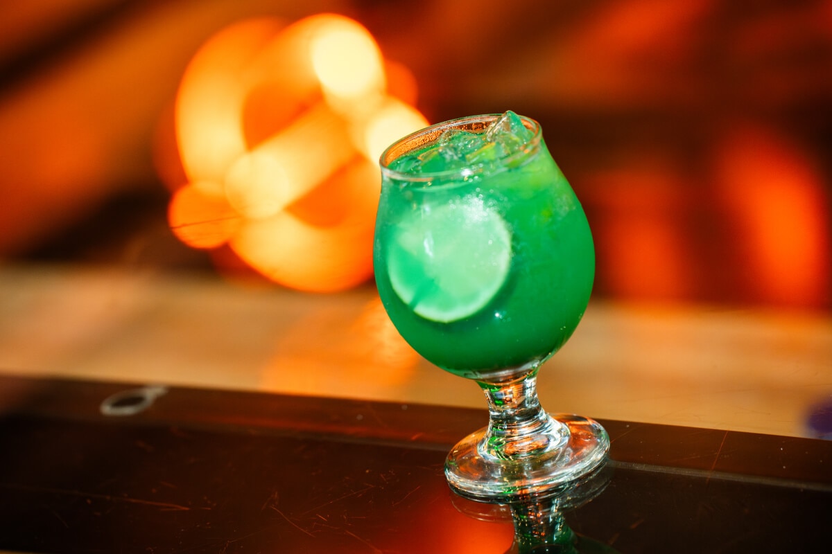  A cocktail glass with green opaque drink and a lime slice on a reflective table with orange lights in the background