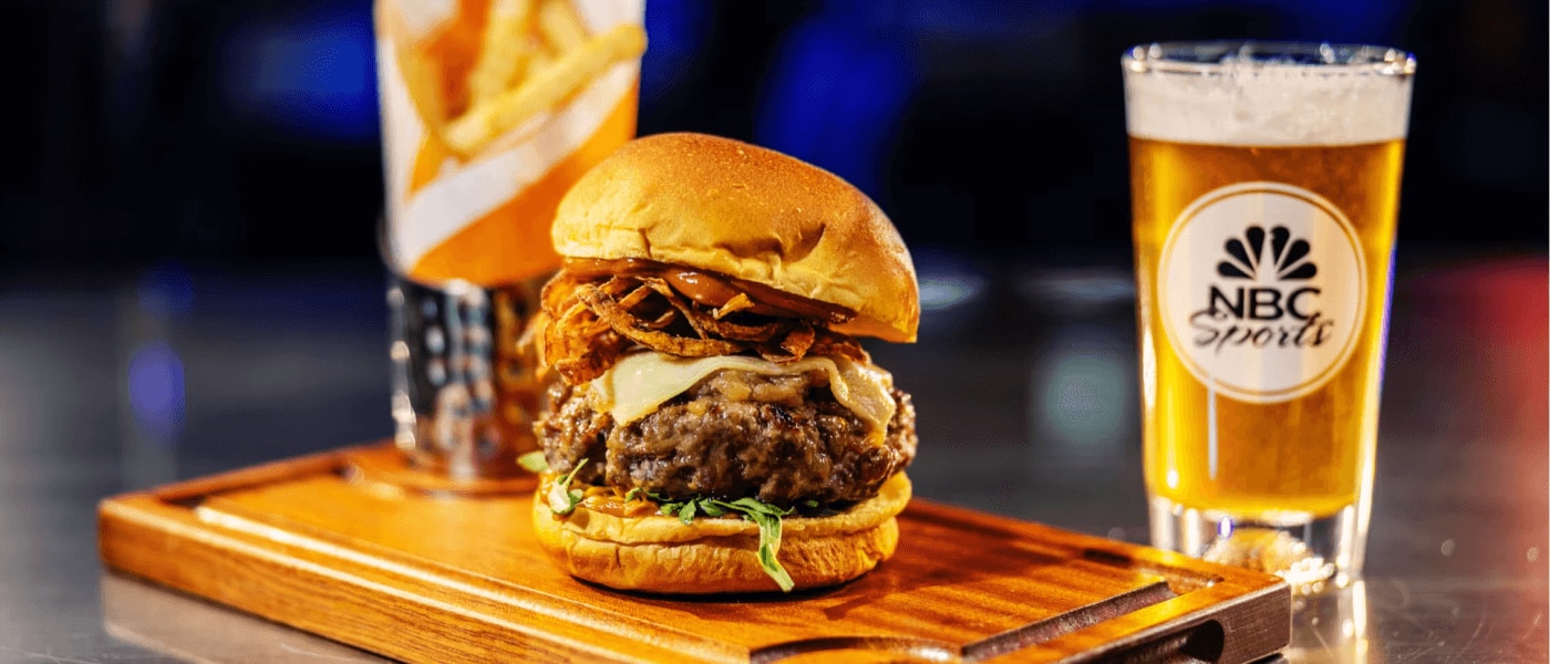 A burger and fries served on a wooden platter next to a beer at NBC Sports Grill & Brew.