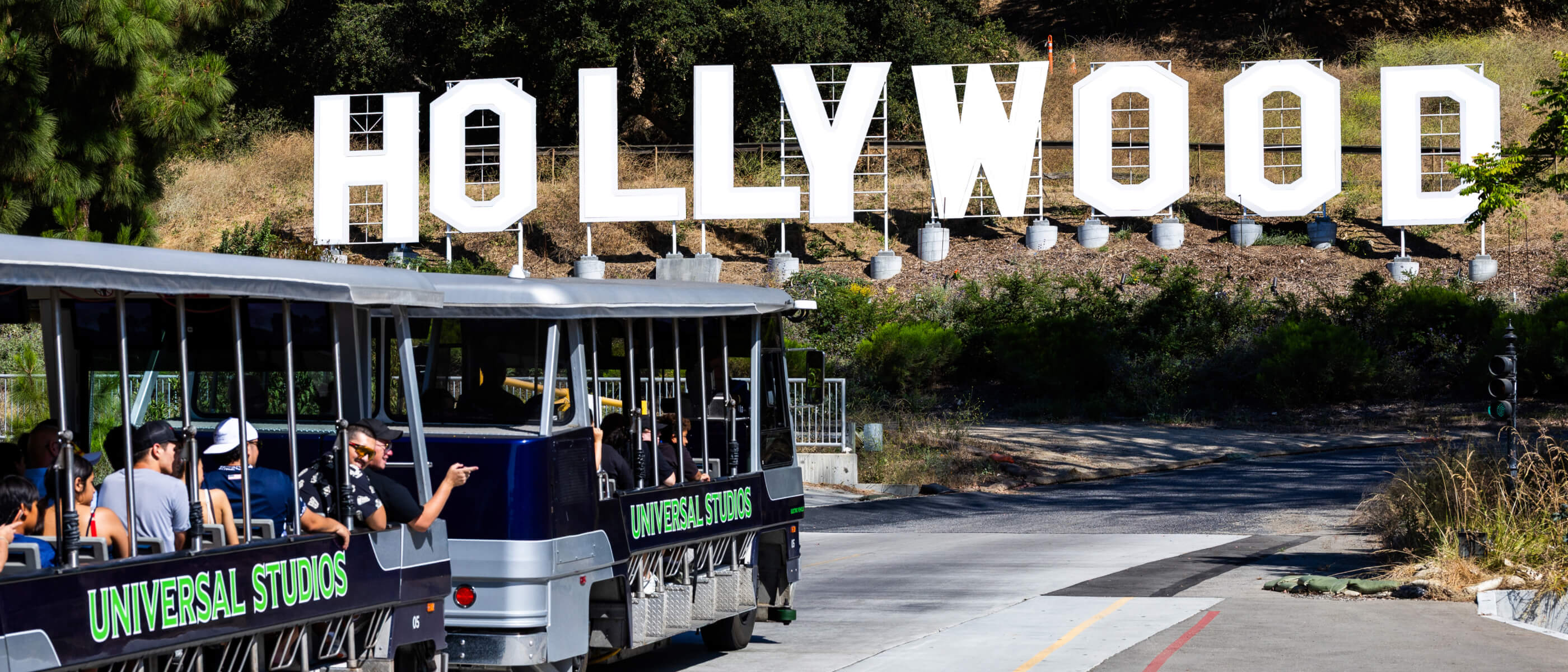 A “UNIVERSAL STUDIOS” tram filled with Guests approaching a large white “HOLLYWOOD” sign on the side of a hill.