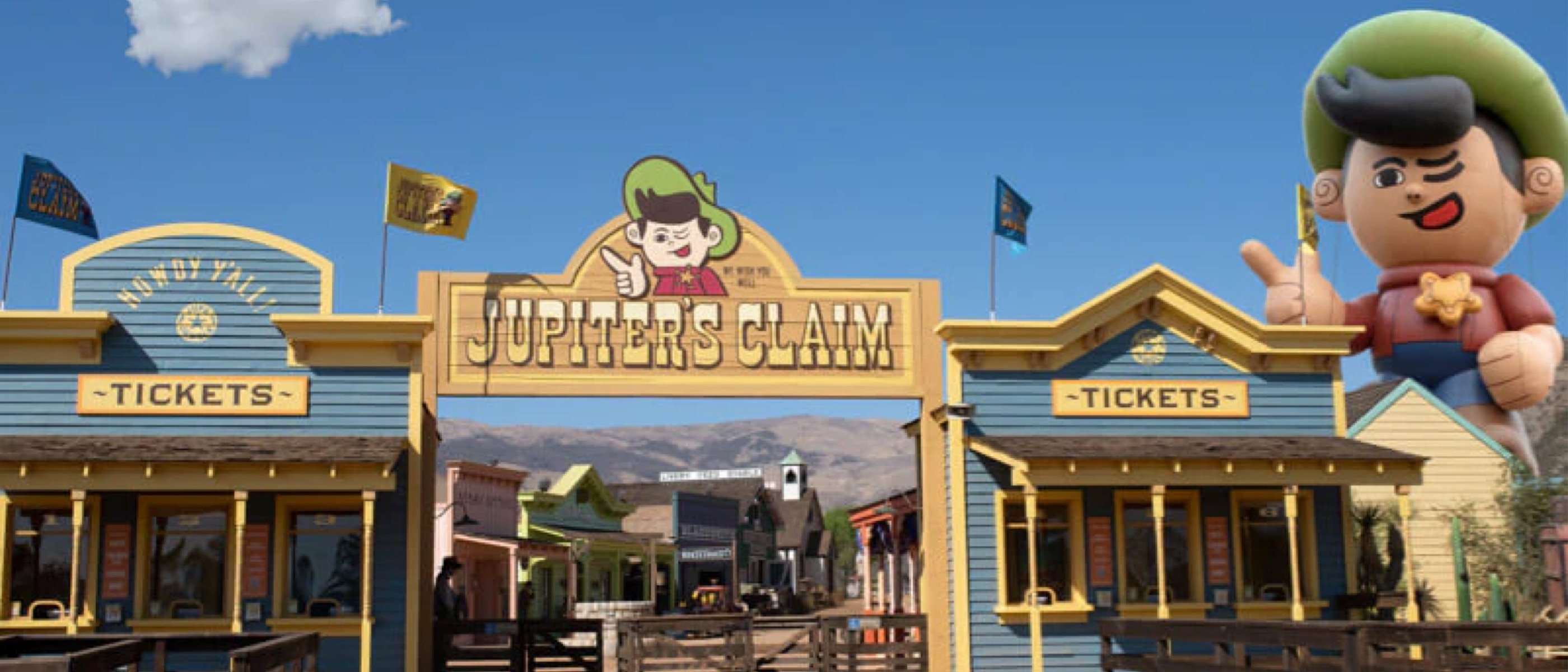 The entrance to a western frontier town tourist attraction features two ticket booths that look like building facades flanking a sign reading "Jupiter's Claim" and a large inflatable cowboy figure towering over the buildings.