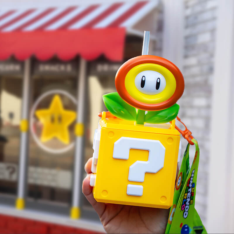A hand holding the SUPER NINTENDO WORLD™ Fire Flower drink bottle in front of the Power Up Cafe.