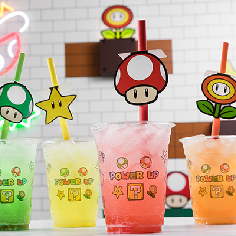 Four clear SUPER NINTENDO WORLD™ POWER UP cups filled with green, yellow, red and orange fizzy drinks.