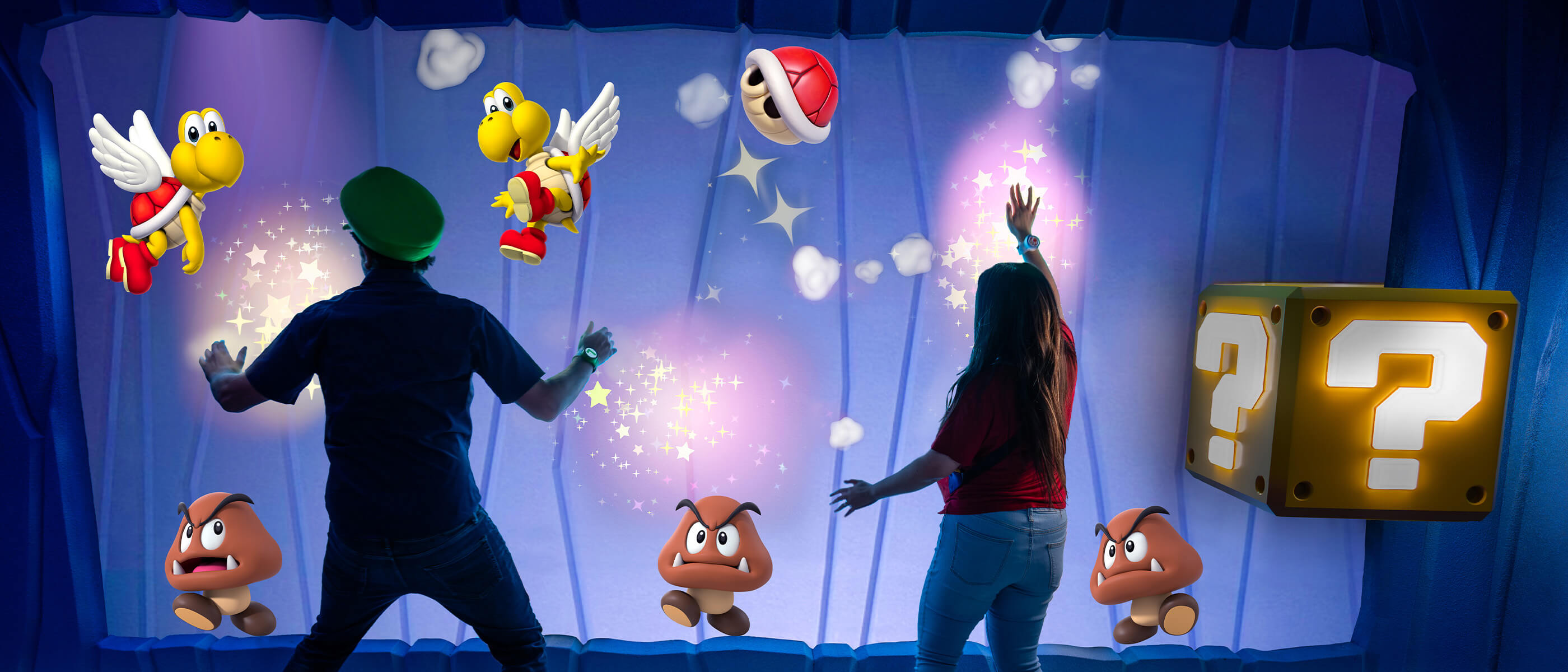 A man in a green Luigi hat and a woman, both wearing Power-Up Bands, swipe at Red Koopa Paratroopas and Goombas on a screen in Frosted Glacier inside SUPER NINTENDO WORLD™ with a question block to the right of the woman.