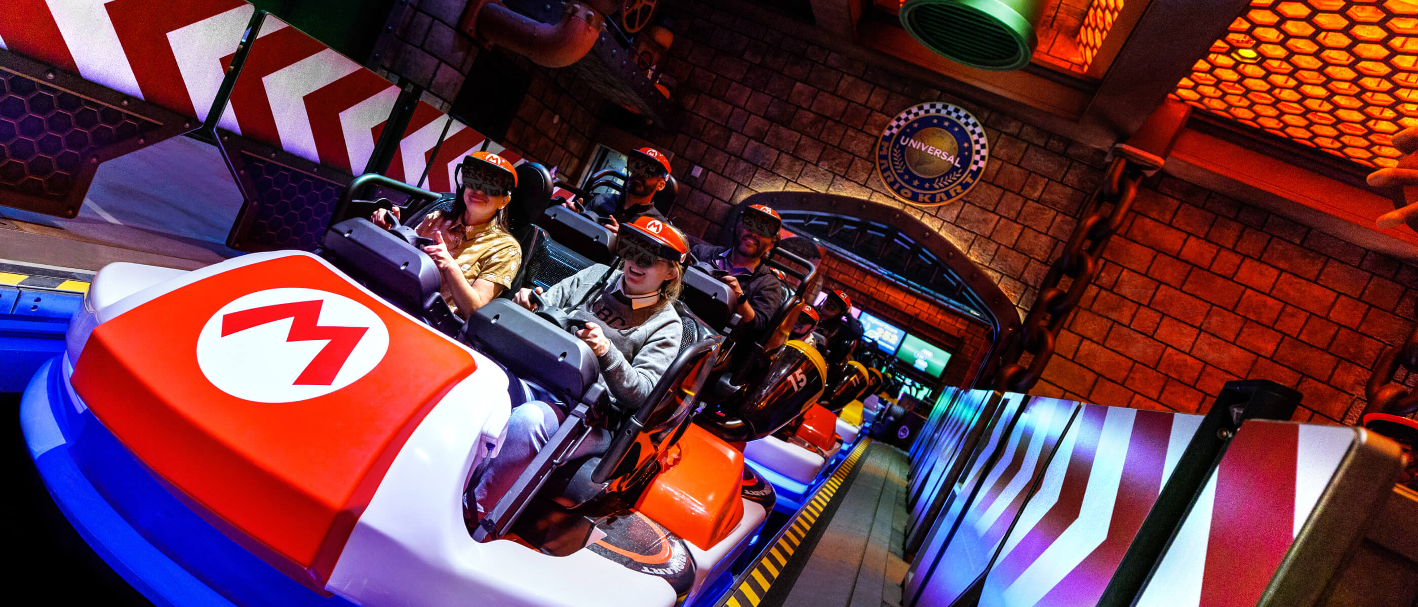 Riders wearing visors and augmented reality goggles sit at their steering wheels in a single line of brightly colored race cart vehicles waiting for the ride to take off.