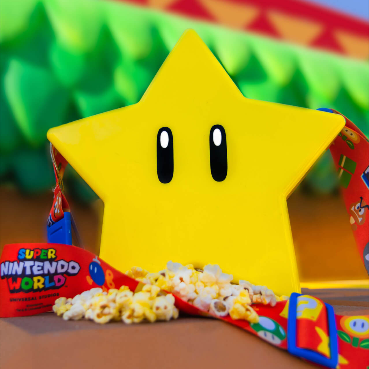 On the left, a popcorn container in the shape of a star with two eyes. The star has a strap attached which reads “SUPER NINTENDO WORLD.” To the right, a Super Mario 1-Up Mushroom drink container. A straw sticks out the top.