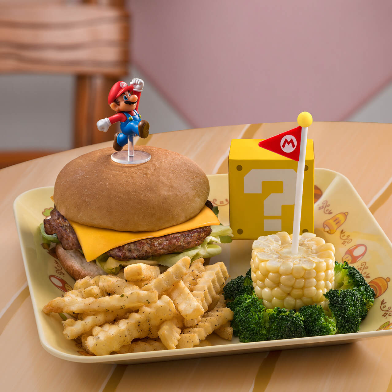 A kids meal consisting of a cheeseburger with a small Mario figure atop the bun; French fries; a section of corn on the cob, surrounded by broccoli, and with a Mario flag sticking from the top; and a square “question box” which holds a cookie inside.