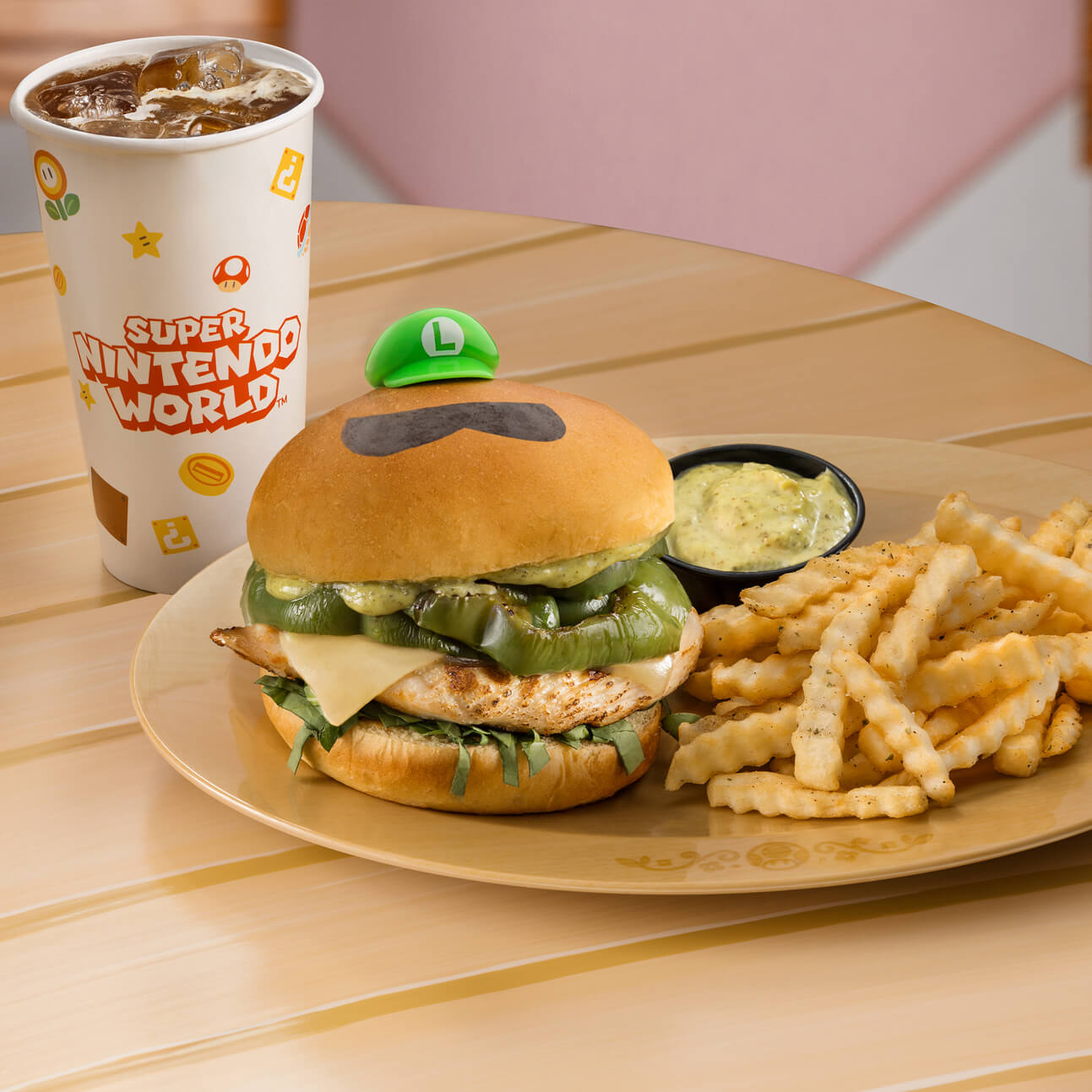 Next to a soft drink cup sits a plate with a grilled chicken sandwich topped with green basil pesto. Atop the bun sits a tiny green Luigi cap and a cartoony mustache. A pile of French fries and a cup of dipping sauce is also on the plate.
