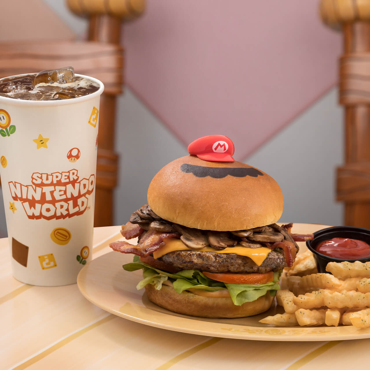 Next to a soft drink cup sits a plate with a cheeseburger topped with mushrooms. Atop the bun sits a tiny red Mario cap and a cartoony mustache. A pile of French fries and a cup of ketchup is also on the plate.