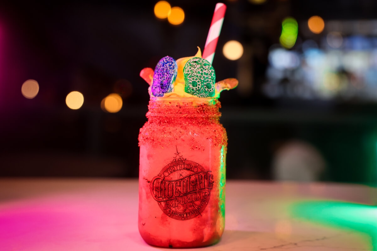A jar-shaped Toothsome Chocolate Emporium glass containing a red drink topped with gummy worms and colored candy skulls.