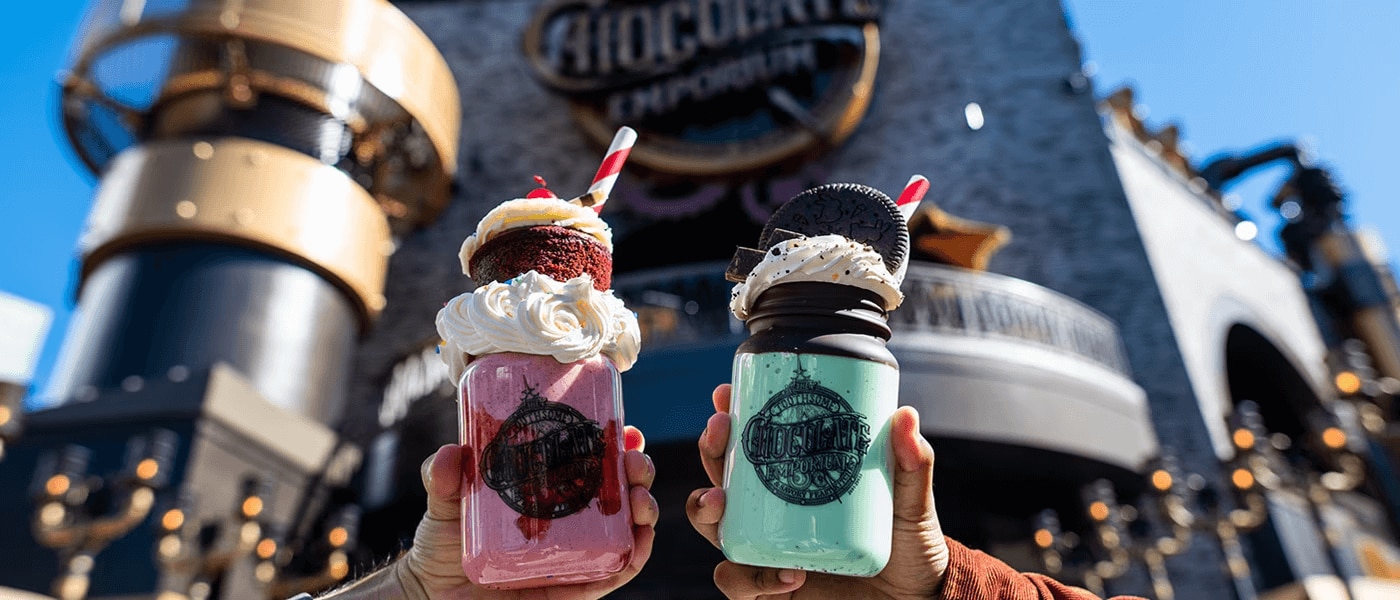 Outside of the Toothsome Chocolate Emporium & Savory Feast Kitchen on Universal CitWalk in Hollywood, two hands hold milkshakes toward the camera. One is a red velvet milkshake with whipped cream and a cupcake and the other is a mint chocolate milkshake with whipped cream and a cookie.