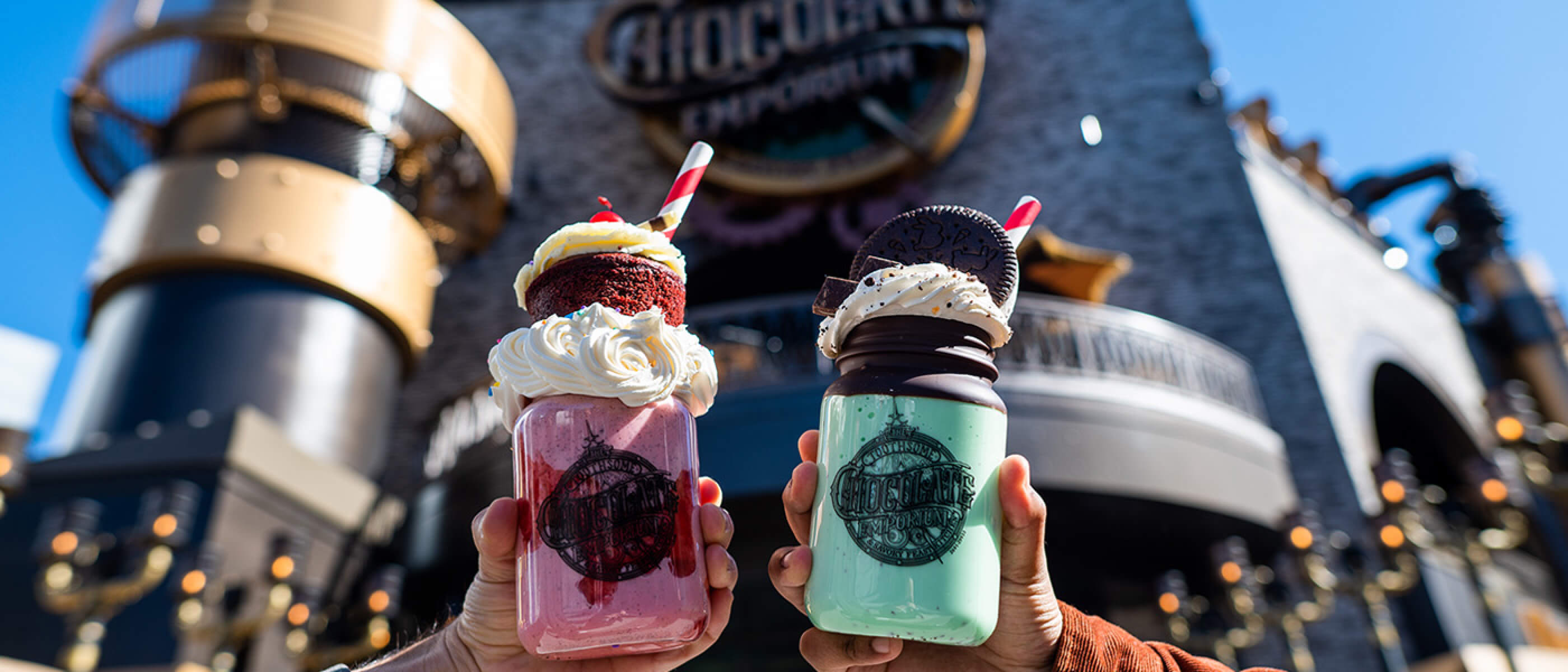 A hand on the left holding a red milkshake next to a hand on the right holding a blue milkshake in front of The Toothsome Chocolate Emporium & Savory Feast Kitchen.