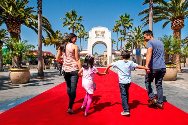 A family standing on a red carpet walks happily towards the arch entrance of Universal Studios Hollywood.