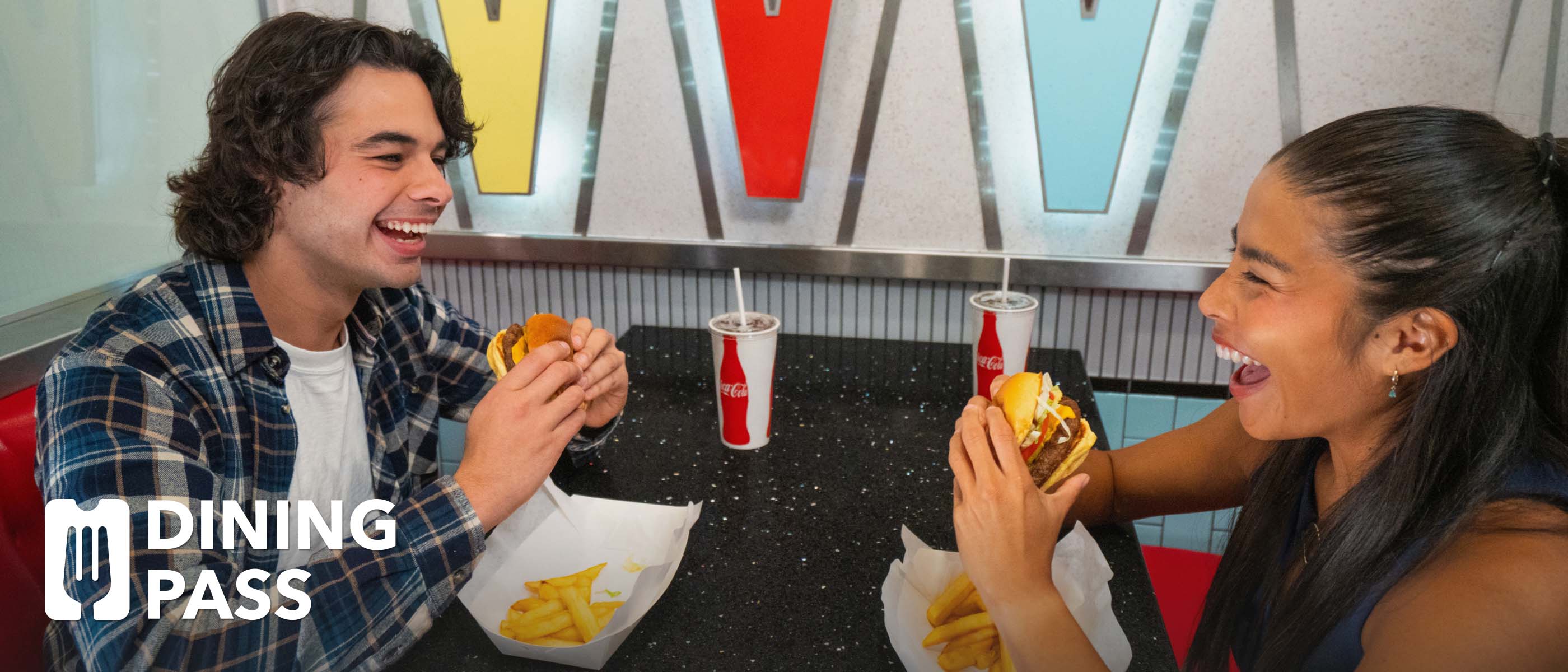 A couple laughing while holding burgers at a table with fries and Coca-Cola fountain drinks in a colorful atmosphere