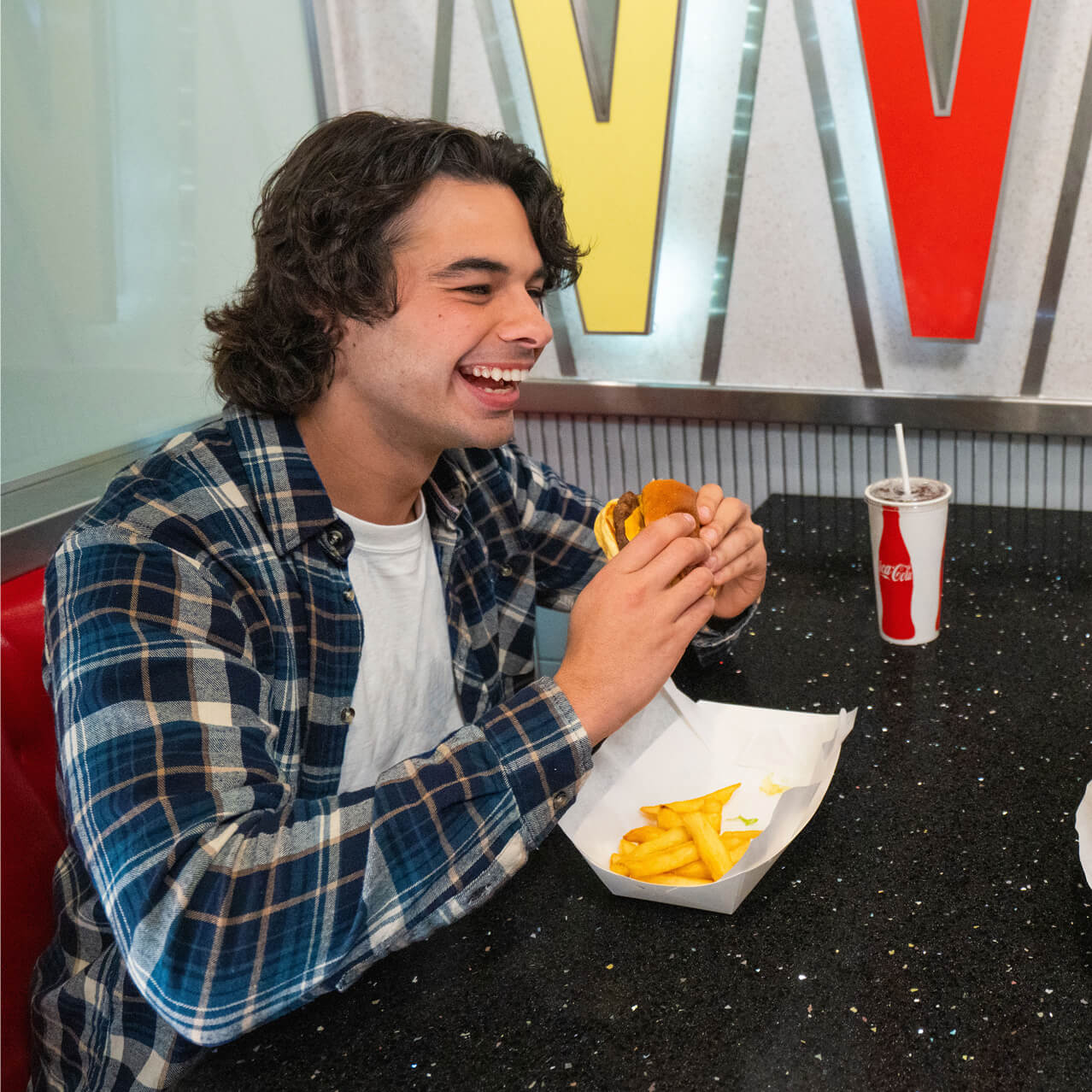 A couple laughing while holding burgers at a table with fries and Coca-Cola fountain drinks in a colorful atmosphere.