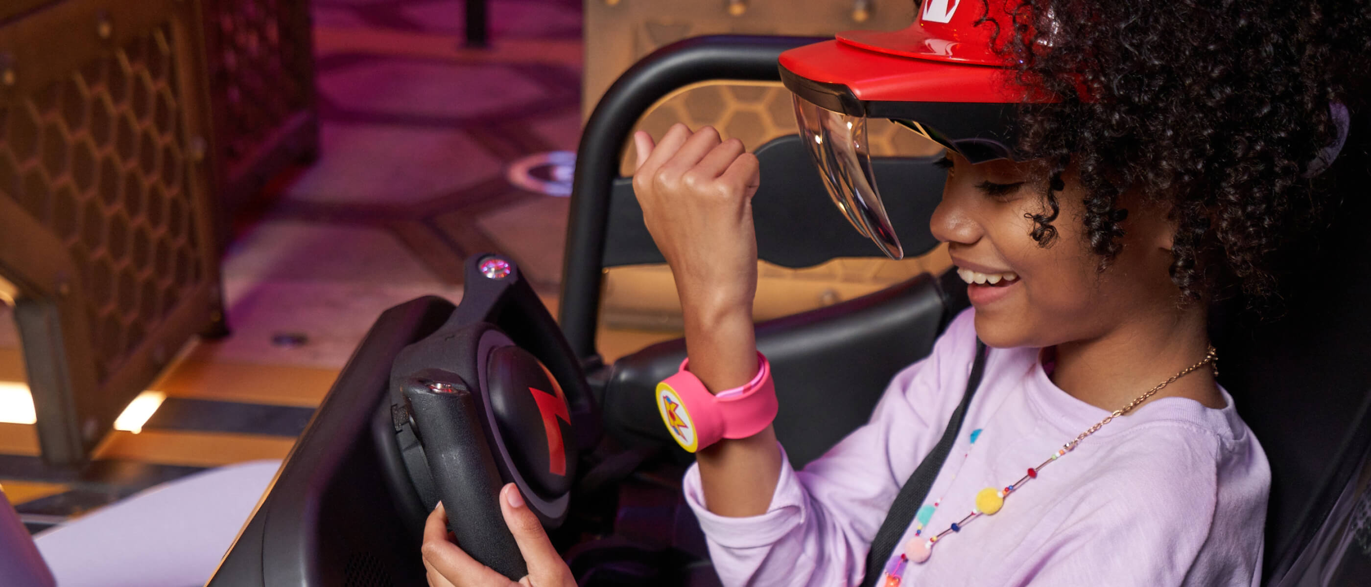 A smiling guest with special goggles tapping her Power-Up Band™ on the steering wheel on Mario Kart™: Bowser’s Challenge.