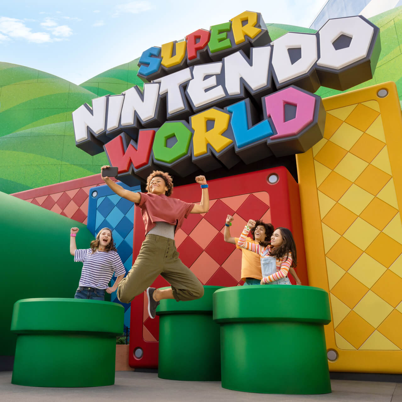 Friends pump their fists in the air while posing for a selfie in front of the SUPER NINTENDO WORLD™ sign.