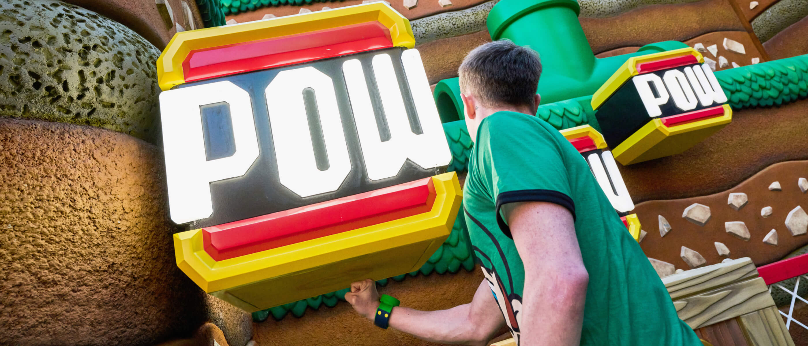 A guest wearing a Power-Up Band™ punches the bottom of a yellow and red block with the word “POW” in SUPER NINTENDO WORLD™.