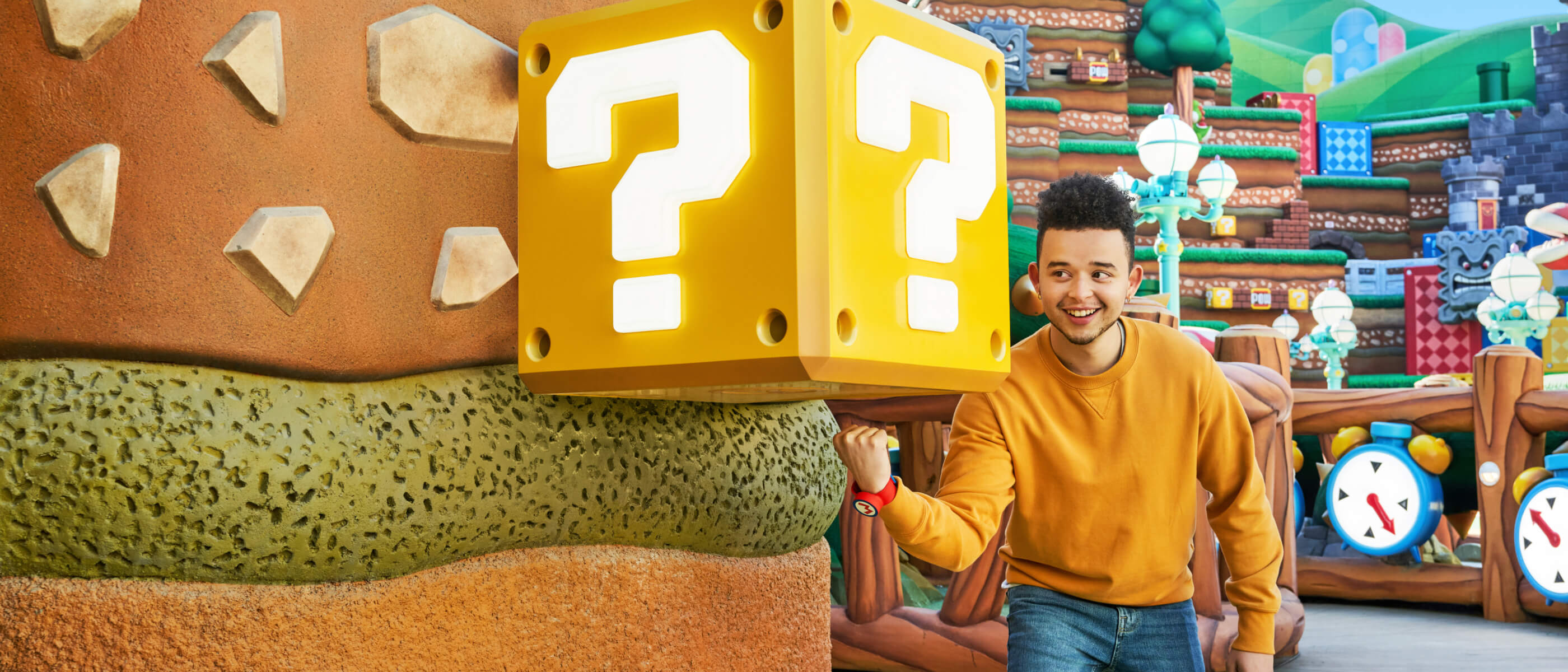 A smiling guest wearing a Power-Up Band™ excitedly punches the bottom of a yellow ? block in SUPER NINTENDO WORLD™.