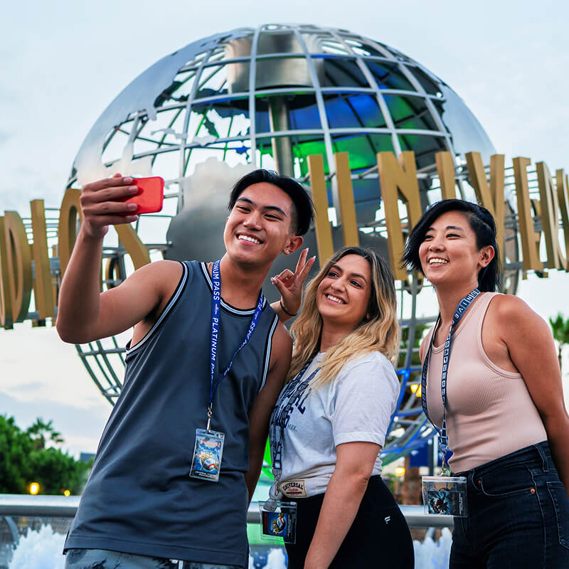 Three smiling friends wearing Pass Member lanyards taking a selfie together in front of the Universal Studios globe.