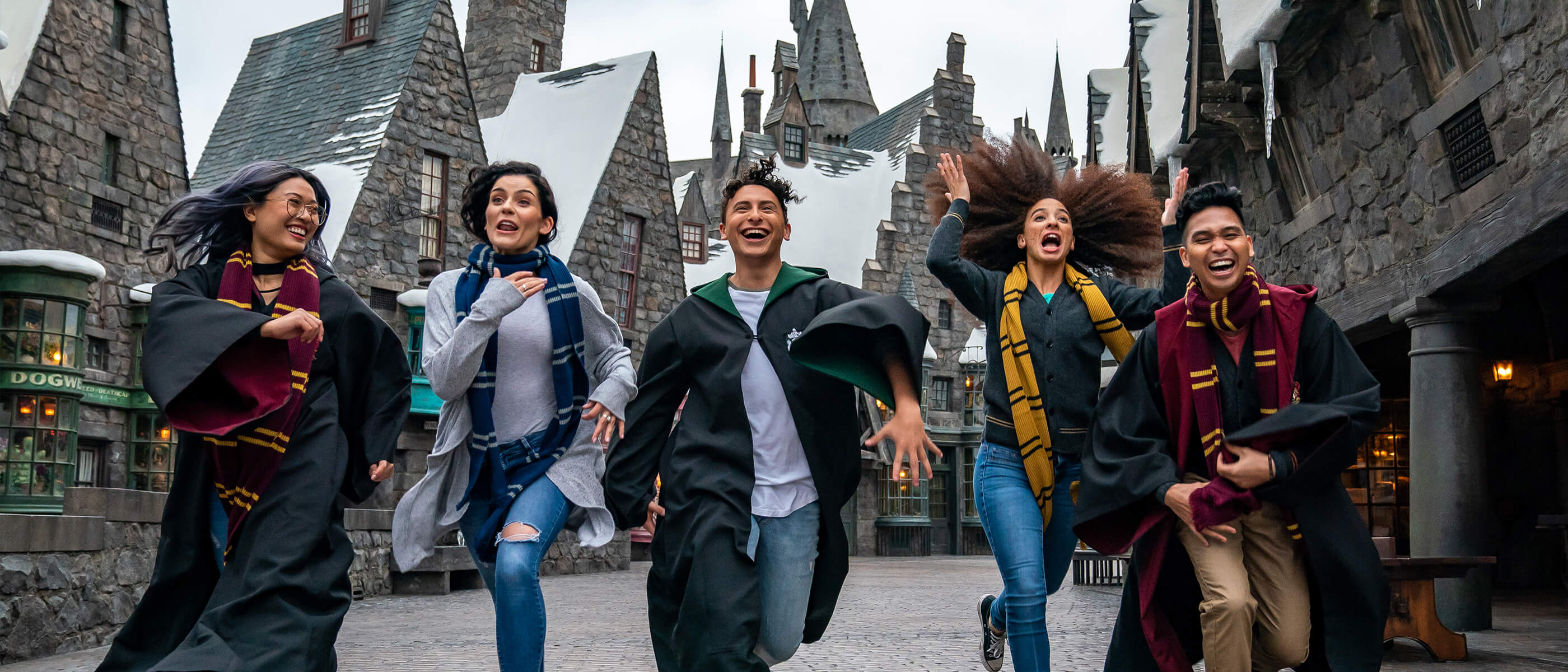 Friends dressed in Hogwarts scarves laugh and run through the streets of Hogsmeade in The Wizarding World of Harry Potter.