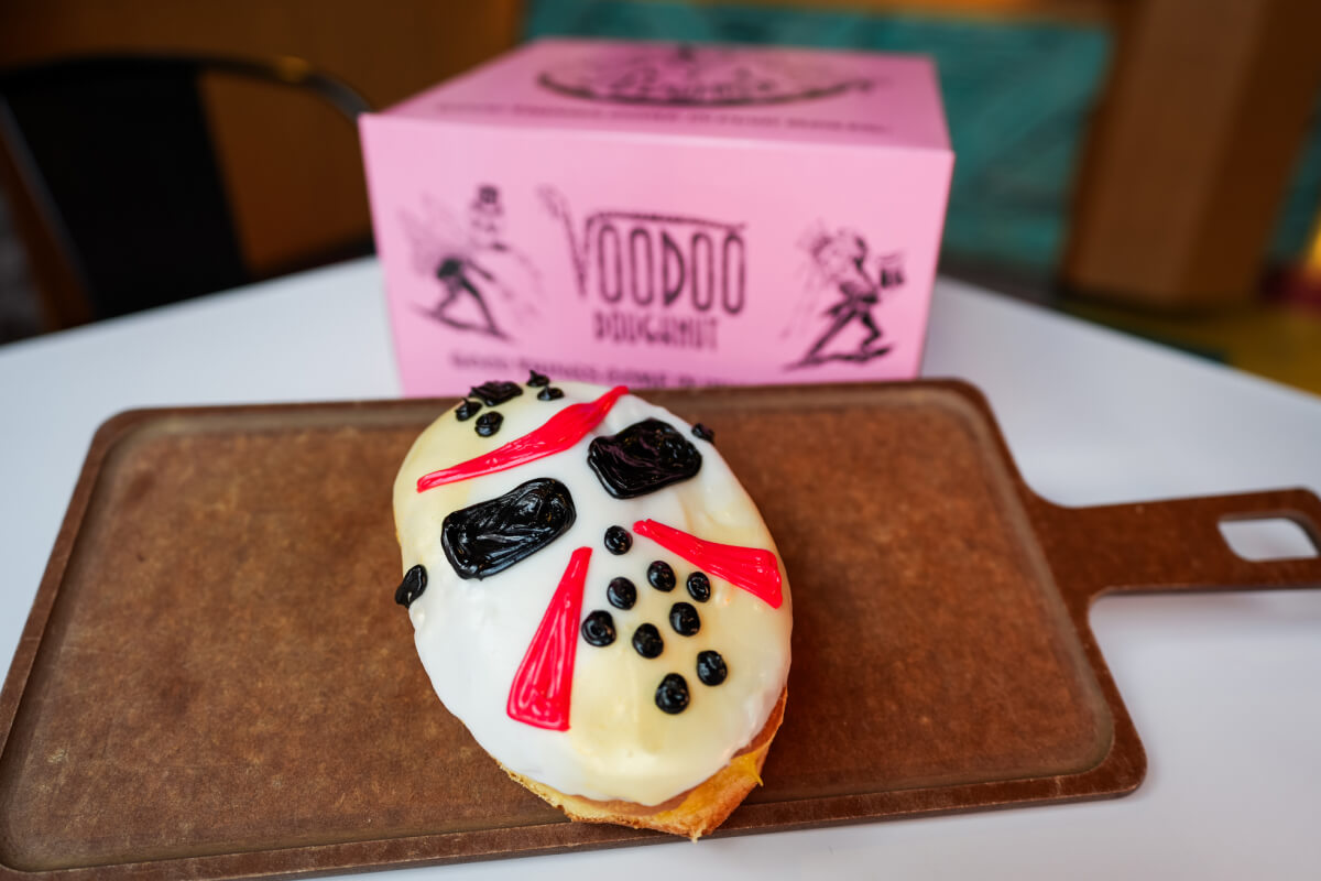 Oval donut with white, black and red frosting resembling a hockey mask, on a wooden tray next to a pink Voodoo Doughnut box.