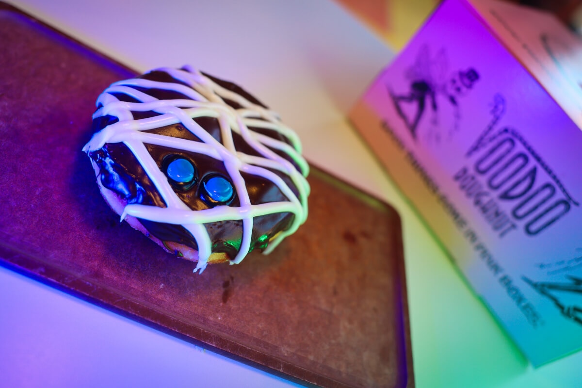 A donut with dark chocolate frosting, strips of white, and two blue dots on a wooden tray next to a pink Voodoo Doughnut box.