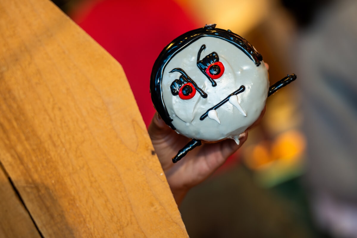 A hand reaching from a coffin holding a doughnut with a vampire face in black, white and red frosting and tiny black wings. 