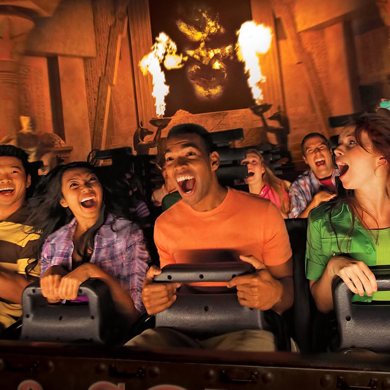 Group of excited passengers riding the Revenge of the Mummy rollercoaster with fire and the face of a mummy behind them