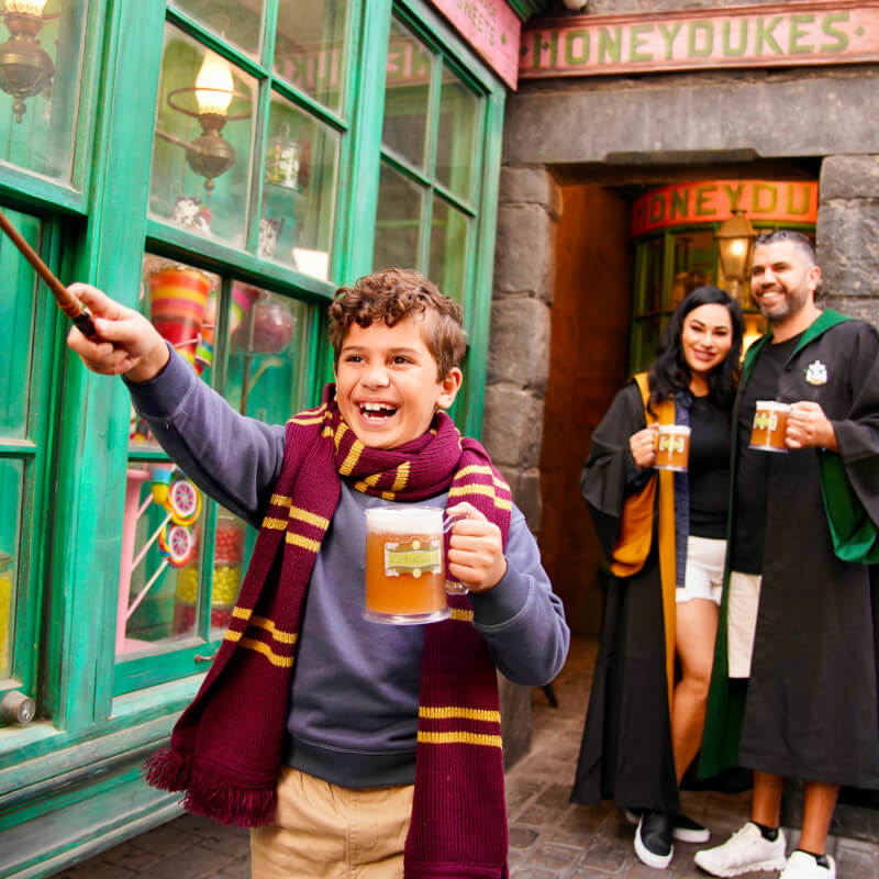 A family of three laugh as they drink Butterbeer™ and the parents look on as their son wields his wand.