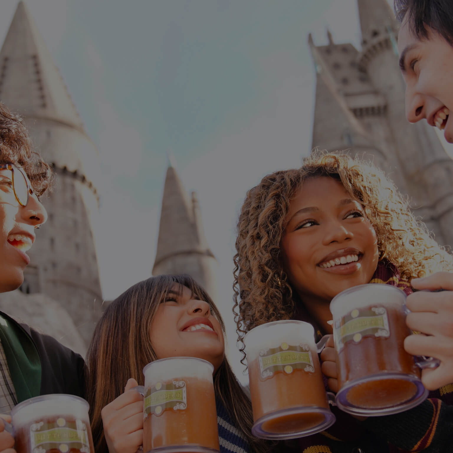 Friends smiling at each other with mugs of Butterbeer™ in front of Hogwarts™ castle.