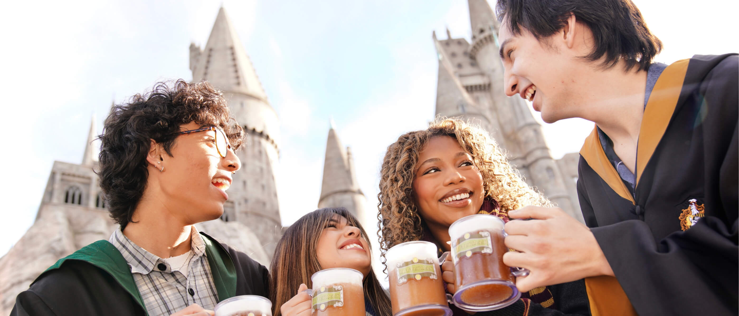 Friends smiling at each other with mugs of Butterbeer™ in front of Hogwarts™ castle.