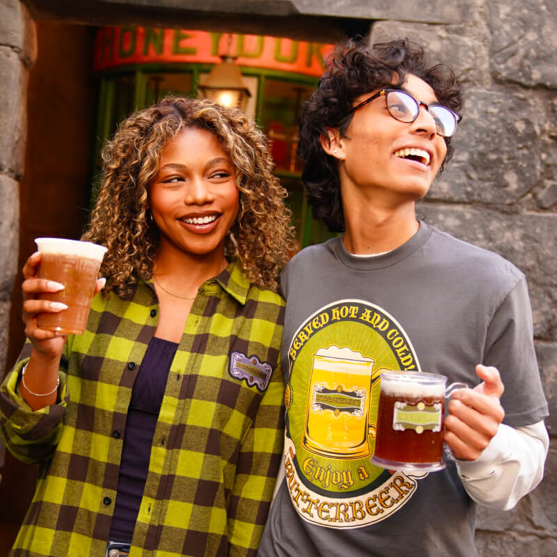 Friends holding Butterbeer™ drinks and wearing a Butterbeer™ flannel and a Butterbeer™ t-shirt in front of Honeydukes™.