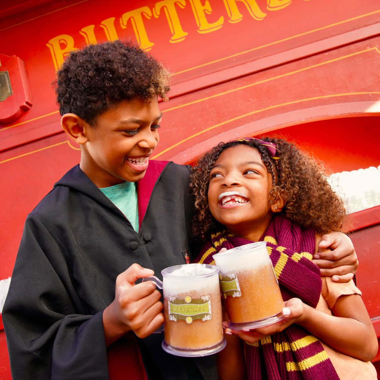 A brother laughs with his sister sporting a frothy foam upper lip as they drink Butterbeer™ in front of a Butterbeer™ cart.