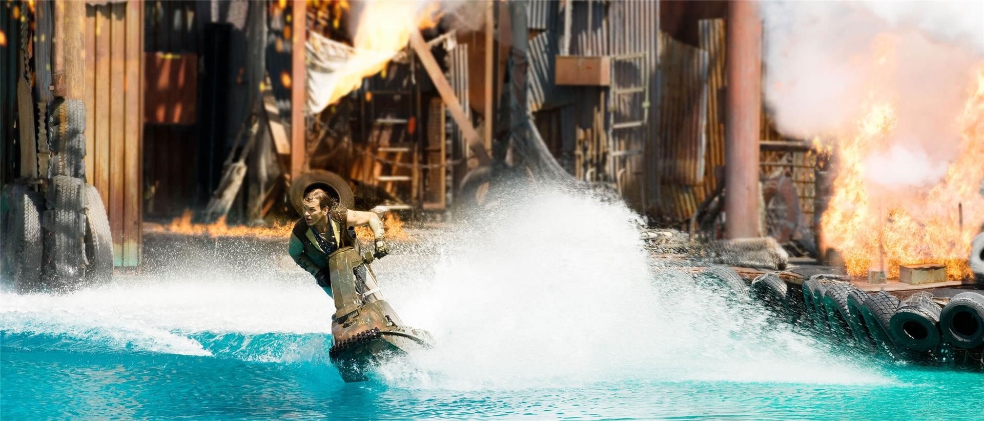 A girl on a jet ski in the water with a building on fire behind.