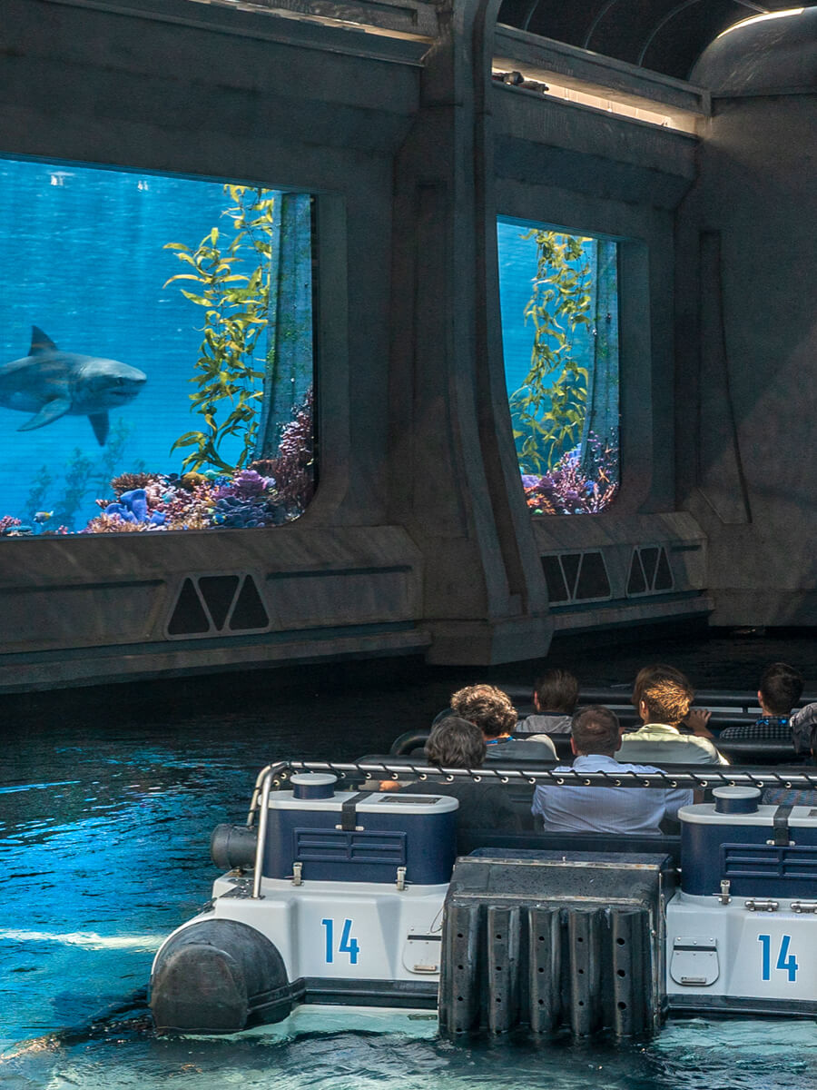 The raft boat from Jurassic World – The Ride moves along the water past aquarium windows where we can see a large shark.