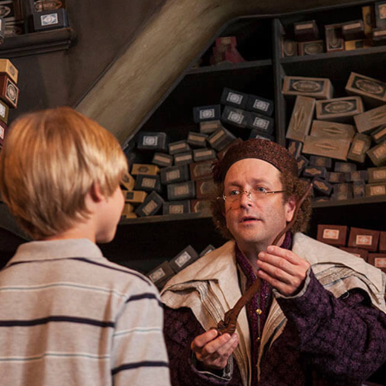 A man dressed up as a wizard in Ollivanders Wand store handing a wand to a young boy.